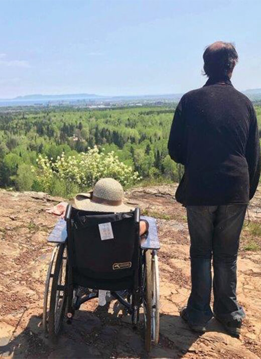 A man and a woman are seen from the back, one next to the other on a mountain overlooking a forest. The man is in a wheelchair, wearing a hat.