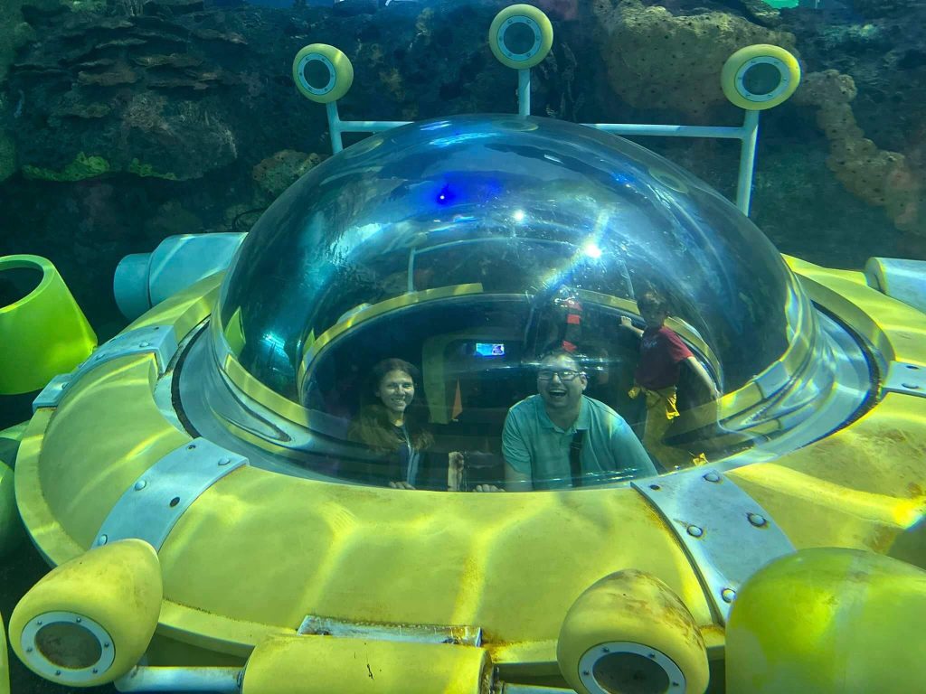 A man and a woman smiling and they're sitting inside a small submarine in an aquarium.
