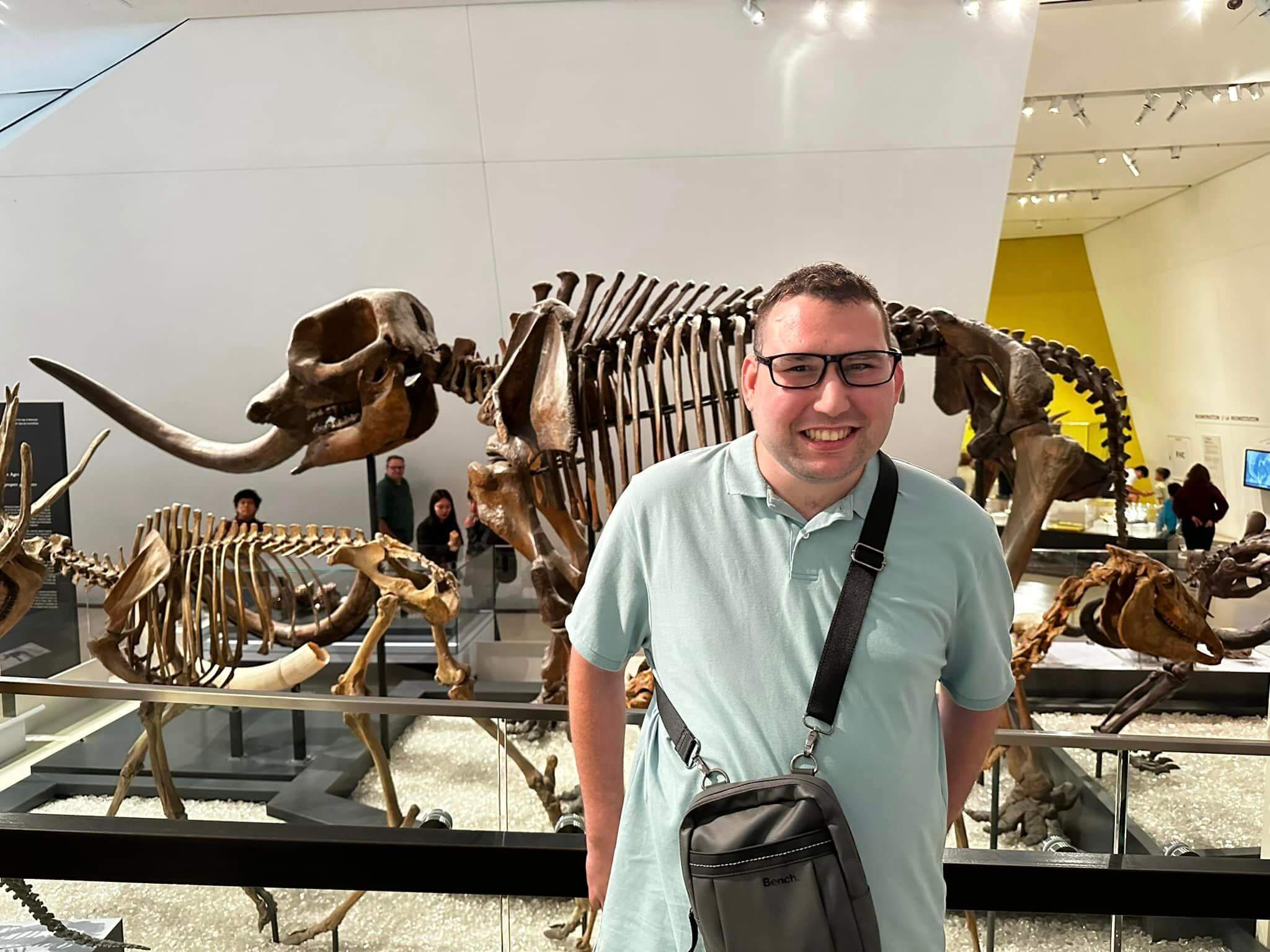 A man is smiling at the camera, and behind him, there is a dinosaur skeleton.