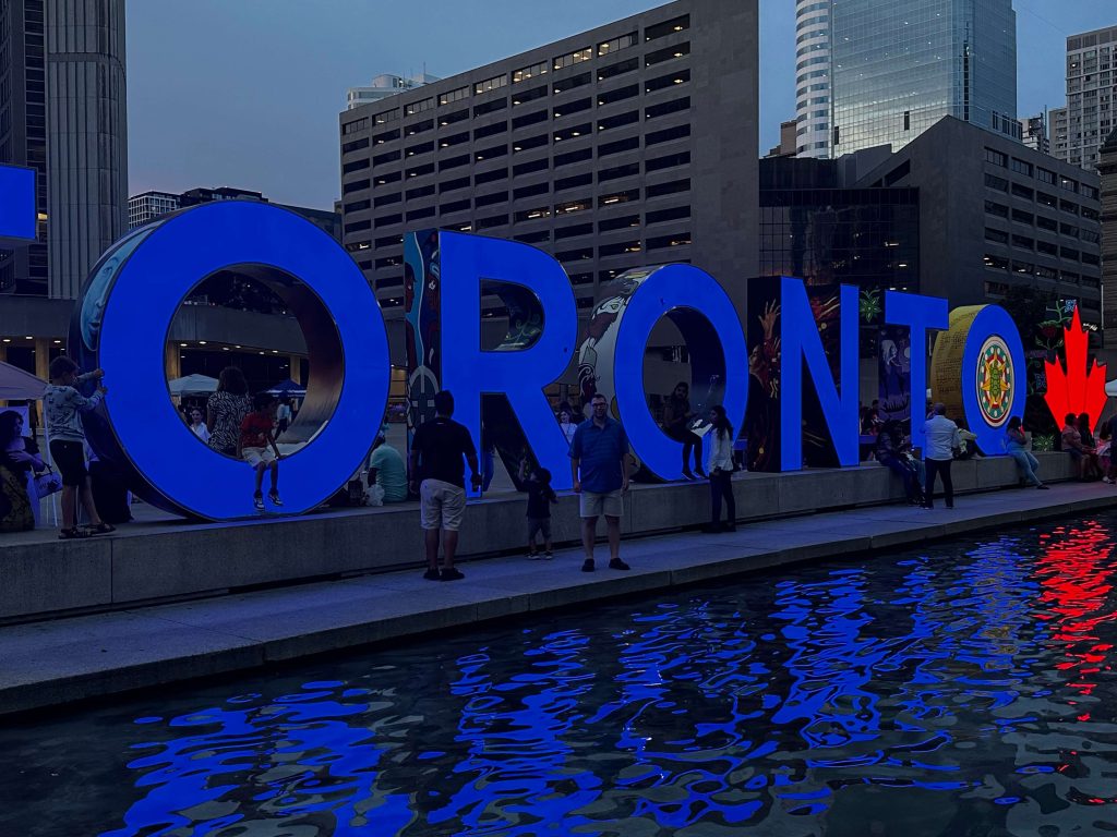A big "TORONTO" sign is above a water fountain. A man is posing for the camera in front of the sign.