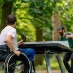 Inclusiveness A disabled man in a wheelchair plays ping pong with an older man in a city park against a backdrop of trees